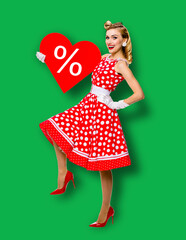 Woman holding red paper heart shape with % sign. Full length body portrait of happy pin up girl. Blond model at retro fashion rockabilly sales ad concept. Green background. Valentine or like symbol.