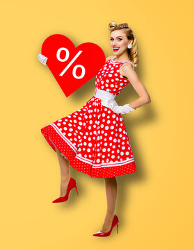 Woman holding red paper heart shape with % sign. Full length body portrait of happy pin up girl. Blond model at retro fashion rockabilly sales ad concept. Yellow background. Valentine or like symbol.