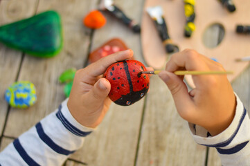 A detailed picture of a child's hands drawing a ladybug on a stone with acrylic paints. Home...