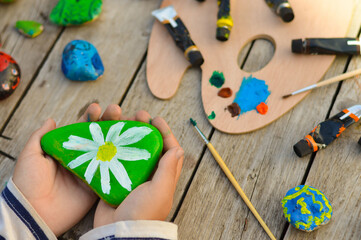 Outdoor workshop in summer. A selective focus of a schoolboy boy holding a painted flat stone in...