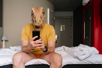 Man with lizard mask using the smartphone in a hotel bed.