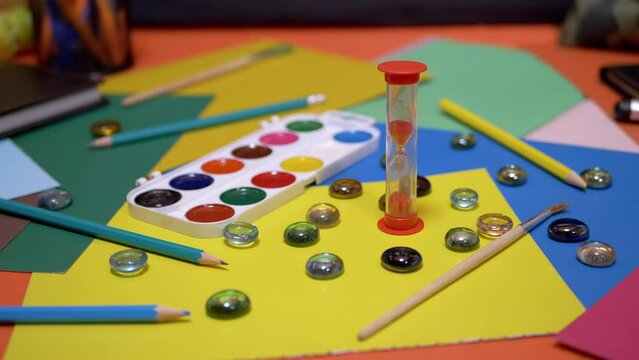 Hourglass on the Background of Scattered Multi Colored Cardboard, School Supplies. Time lapse. Time concept. Flowing sand. End of lesson. Creativity, homework. Yellow background. Childhood. Education.