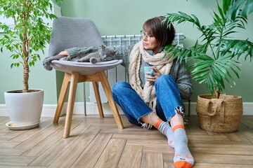 Cold winter season, woman with cat on chair near heating radiator