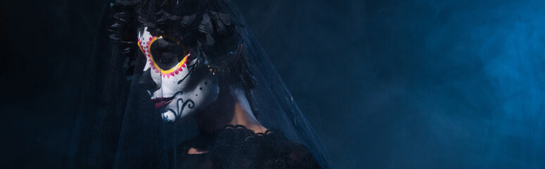 woman in catrina makeup and black wreath with veil on dark blue foggy background, banner.
