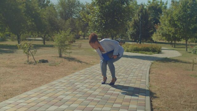 Elegant attractive black female with documents and tablet pc, wearing business casual clothes and high heels twisting ankle during walk outdoors and touching injured leg with painful expression.