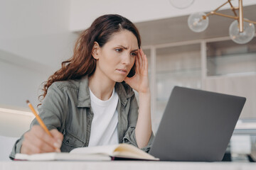 Frowning female student learning online at laptop, taking notes, sitting at desk at home. E-learning