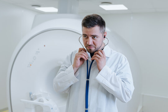A male doctor in a white coat with a stethoscope in his hands against the background of a modern CT scanner in the clinic.