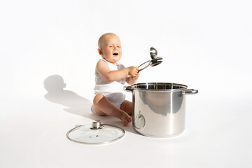 Little cute baby chef holding spoon sitting near big cooking pot with kitchen, utensils, accessories on white background. Child cook, food boy. Copy space