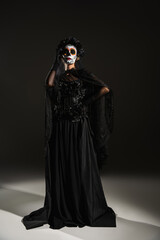 full length of woman in black witch dress and scary halloween makeup posing with hand on hip on dark background.