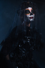 woman in traditional santa muerte makeup and black lace veil looking at camera on dark blue background.