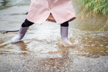 Little girl in pink waterproof raincoat, purple rubber boots funny jumps through puddles on street road in rainy day weather. Spring, autumn. Children's fun after rain. Outdoors recreation, activity