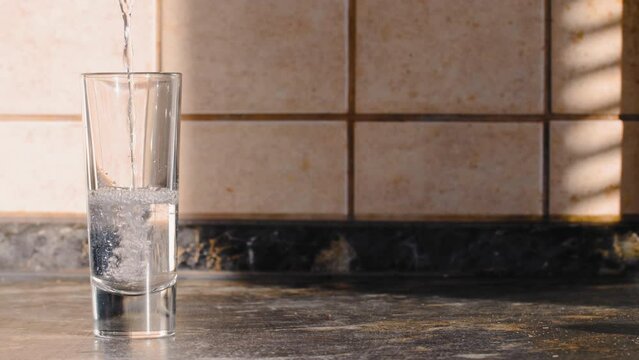 Liquid is poured into a glass. Cold Vodka is poured into a tall glass on a table against a background of light tiles in the kitchen. Transparent alcohol drink. Pure water with bubbles. Close-up