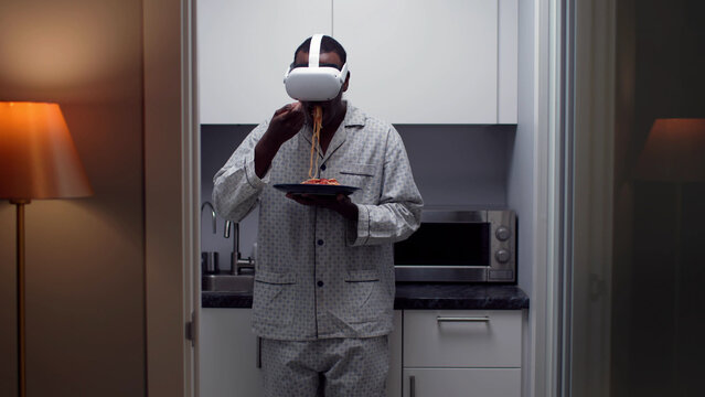 African-American man in pajamas eat pasta in vr goggles standing in kitchen