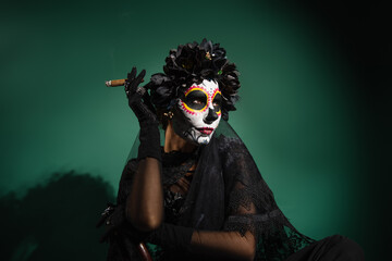 Woman in santa muerte halloween costume holding cigar on green background.
