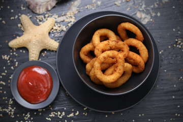 Deep fried squid rings served with tomato sauce, ketchup. Panko calamari in a black bowl, on a table, top view.