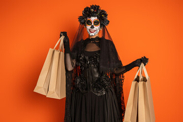 Obraz premium joyful woman in traditional santa muerte makeup and black costume standing with shopping bags isolated on orange.