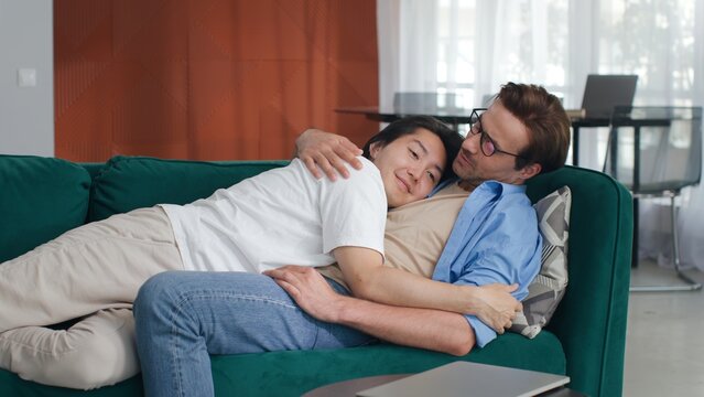 Diverse attractive male gay couple relax together on sofa at home. Boyfriend puts his hand on partner's and they hug. They are happy and smiling. They are casually dressed and room has modern interior