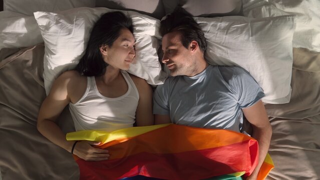 Top view of male gay couple lying in bed and covering with rainbow flag