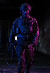 Army soldier in Combat Uniforms with an assault rifle and combat helmet night mission dark background. Blue and purple gel light effect. 
