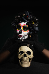 Woman in mexican day of death costume looking at camera near skull isolated on black.