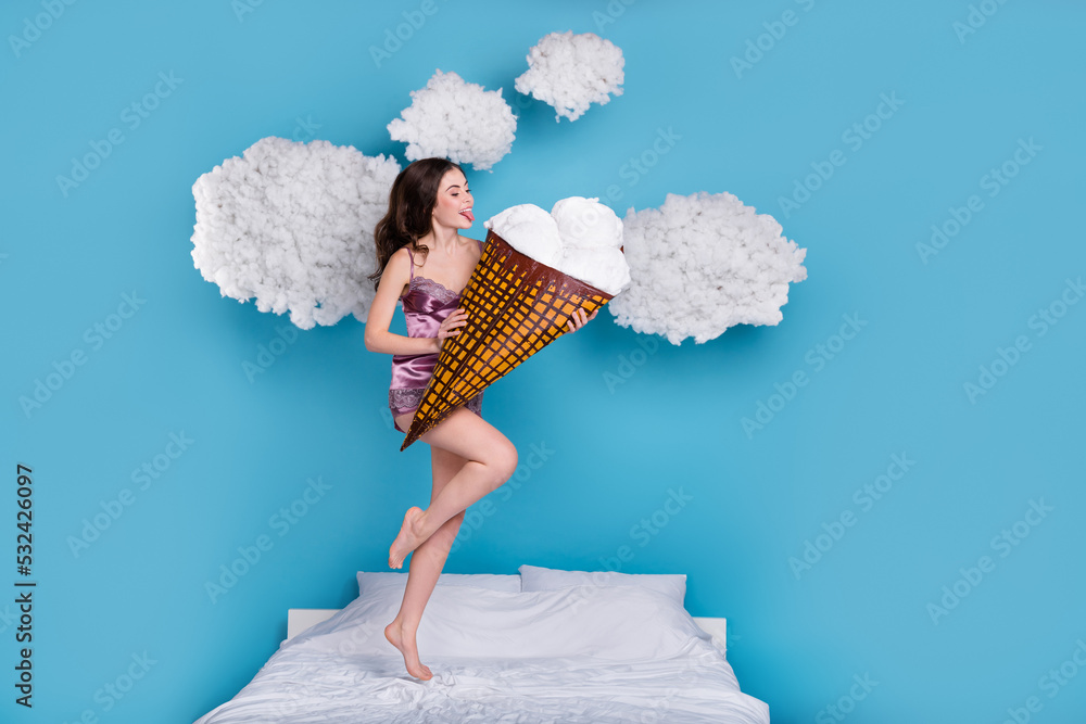 Sticker Full length body size view of beautiful trendy girl jumping on bed licking cone gelato isolated over bright blue color background