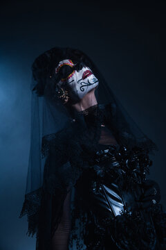 woman in creepy santa muerte makeup and black costume posing with closed eyes on black and blue background.