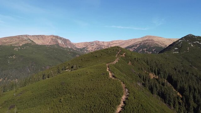 Aerial footage of a mountain ridge with a path between green forest and bare mountain peaks under the blue sky on the background