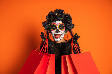 smiling woman in black halloween costume and sugar skull makeup holding shopping bags and looking at camera on orange background.