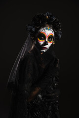 woman in traditional santa muerte makeup and dark costume with wreath looking at camera isolated on black.