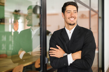 Ambitious hispanic man in formal wear stands with arms crossed in contemporary office space and looks away with smile, portrait of purposeful latin businessman, ceo indoor. Diversity of office staff