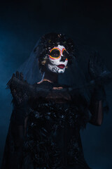 woman in witch costume and sugar skull makeup holding lace veil on dark blue background.