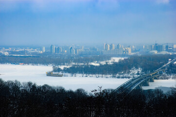 Fototapeta na wymiar Panoramic view of the Kiev's left bank during a foggy winter day. The Dnieper river is frozen. Trees, road, bridge and buildings appear in the distance