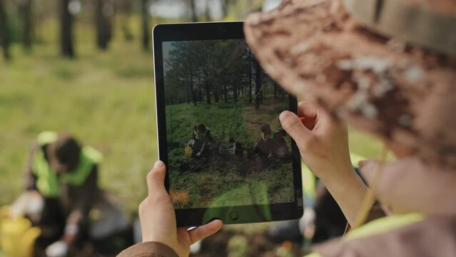 Over-shoulder of unrecognizable person using tablet computer, taking pictures of gardeners planting in forest on sunny day