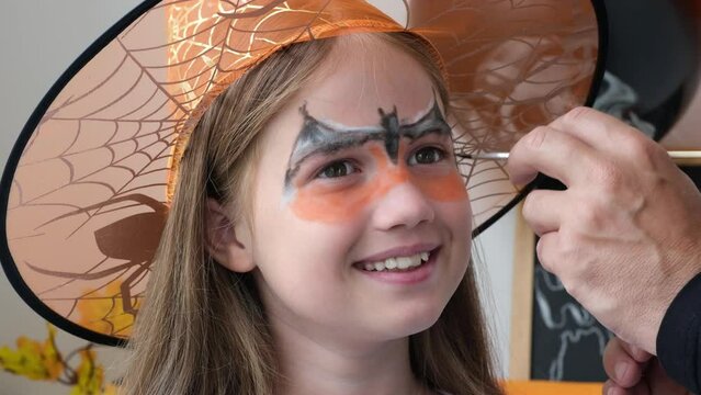 Happy family preparing for Halloween. Man hand draws aqua makeup on kid girl face for holiday party. Dad and daughter getting ready for halloween making make-up. Close-up