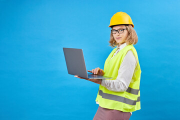 Studio shot of female professional engineering foreman manager wear hard helmet and reflective safety vest standing and smiling, holding laptop computer in studio on blue background.