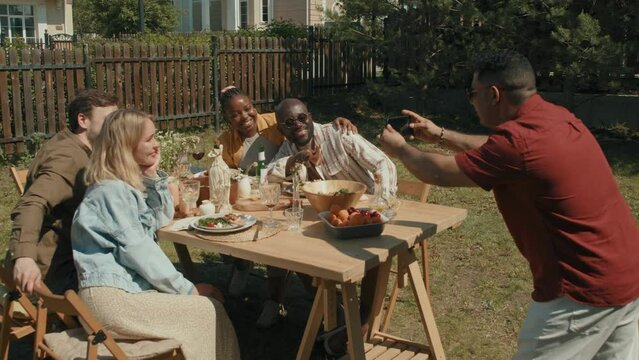 Middle Eastern man taking group photo of his friends sitting together at dining table outdoors on summer day using smartphone camera