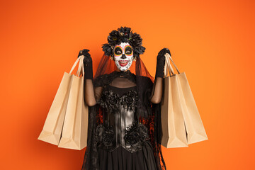 Obraz premium excited woman in black halloween costume with wreath and scary makeup holding shopping bags isolated on orange.