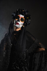 woman in halloween makeup and witch costume with black lace veil standing with hand on waist on dark background.