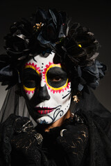 Portrait of woman in wreath and catrina makeup looking at camera on black background.