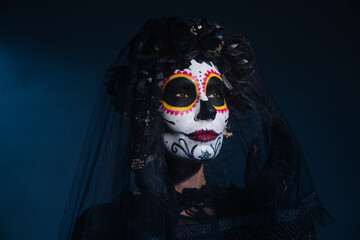 woman in traditional mexican catrina makeup touching veil and looking away on dark blue background.