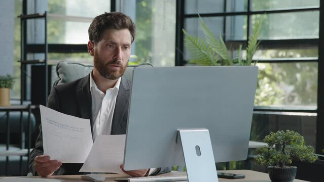 Caucasian pensive bearded man boss manager adult 40s businessman sitting in office busy with paperwork and computer check data in papers search information look at screen corporate company app project