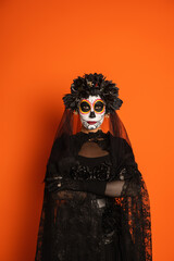 woman in black witch costume and traditional mexican day of dead makeup standing with crossed arms isolated on orange, banner.