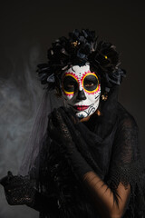 woman in black wreath and traditional santa muerte makeup looking at camera on dark foggy background.