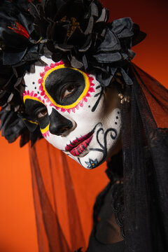 portrait of woman in traditional mexican day of dead makeup and black wreath looking at camera on orange background.