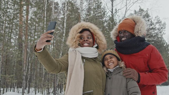 Happy African American family in warm winterwear taking selfie photos in forest park using smartphone