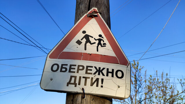 Warning road sign Caution children with some minor damage, attached to a telegraph pole with an inscription in Ukrainian language "Caution children!", wires and tree branches in the background