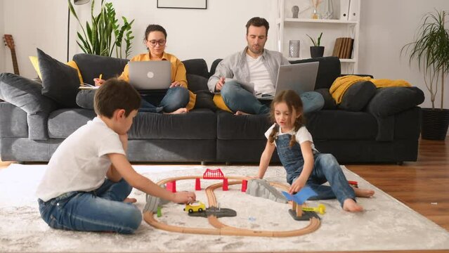 Busy parents work remotely with laptops sitting on the couch in living room, two sweet kids playing with toys on the foreground, freelancer parents working on the distance during quarantine