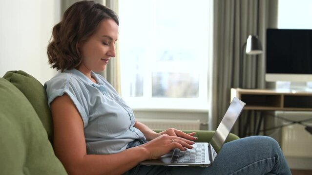 Young freelancer female employee sitting on the sofa with the laptop, working online, concentrated woman typing