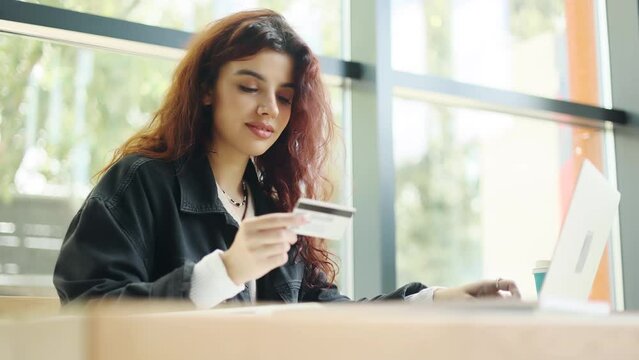 Young attractive curly woman paying with credit card on laptop sitting in a light cafe. Happy customer. High quality 4k footage