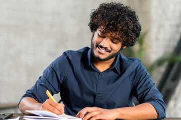 Online training. Young guy student with curly hair studying at home while sitting in the modern room. He writing with a pen in a notebook - obrazy, fototapety, plakaty